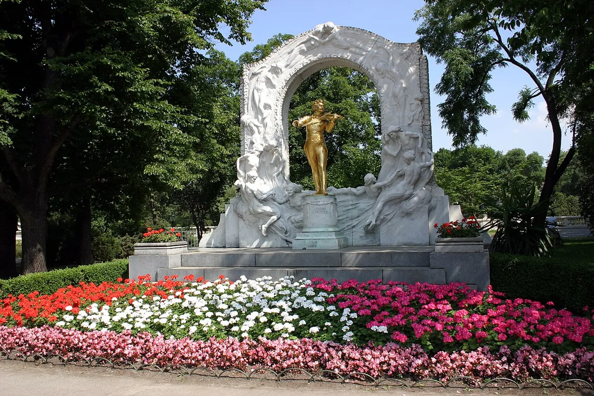 The gilded statue of Johann Strauss II playing the violin under a white marble arch in Vienna's Stadtpark, with flower beds in front